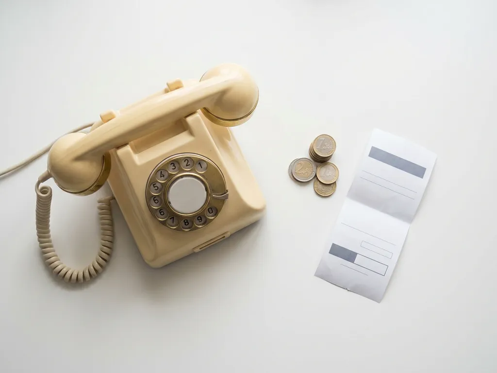 Vintage draaitelefoon op wit bureau naast gestapelde euromunten en een bon, symbool voor telefoonbereikbaarheid en inkomsten.