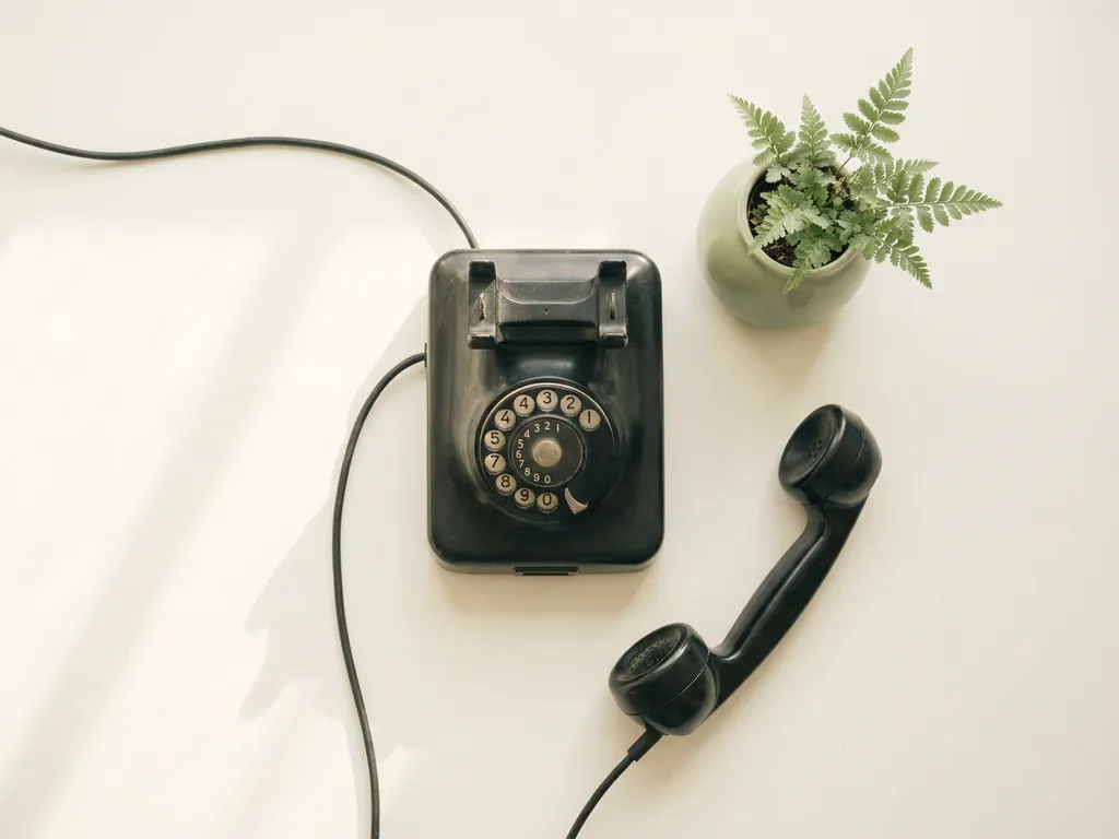 Vintage zwarte telefoonhoorn naast de haak op wit oppervlak, met klein groen potplantje en zachte ochtendschaduwen, bovenaanzicht.