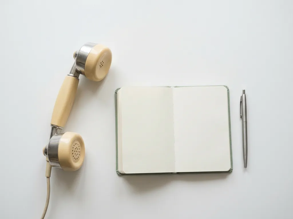 Vintage telefoonhoorn op wit oppervlak naast een open notitieboekje met pen, minimalistische flatlay in zilver en ivoor tinten.